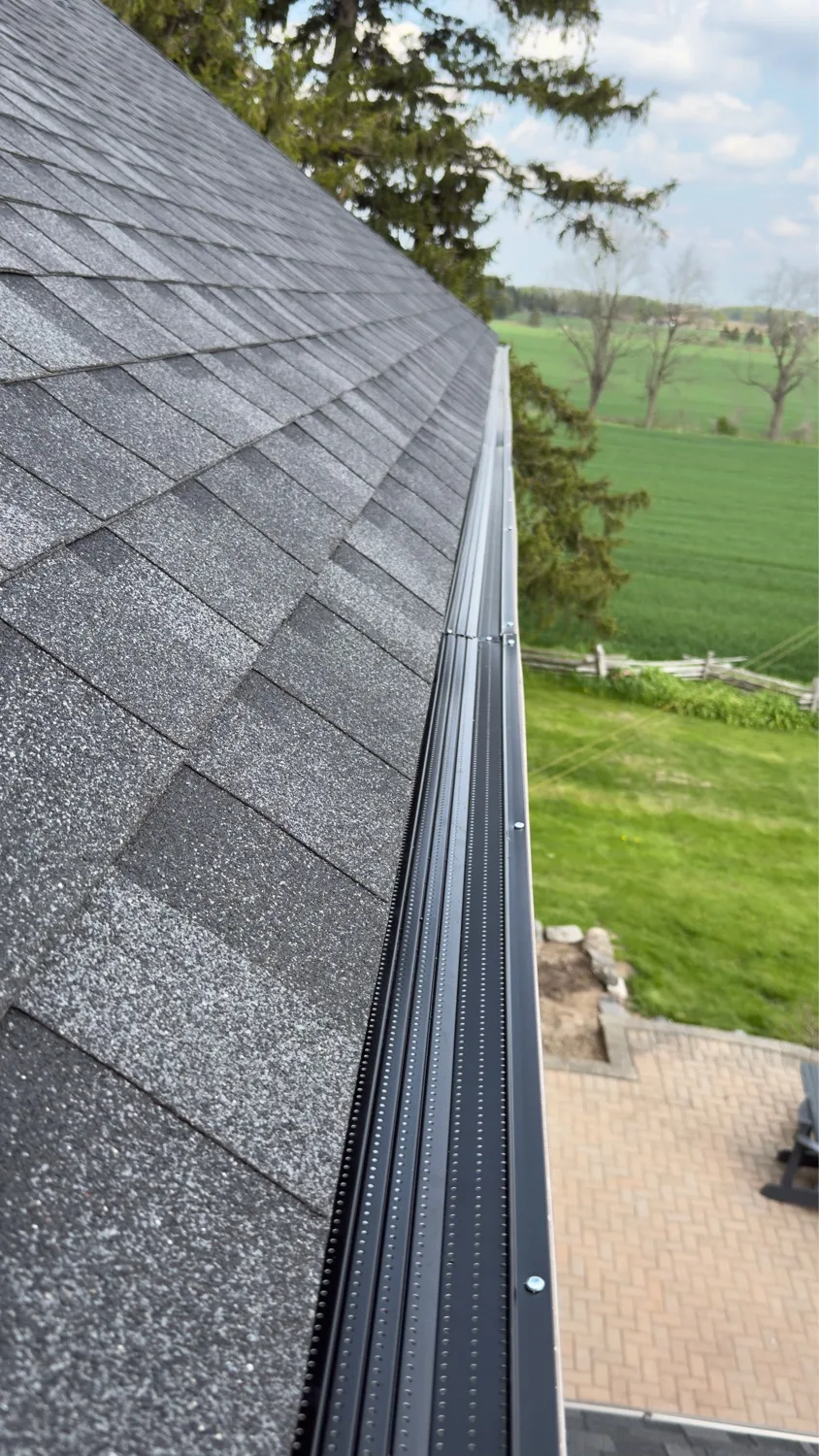 Black gutter guard along grey asphalt shingle roof with rural Waterloo fields in background