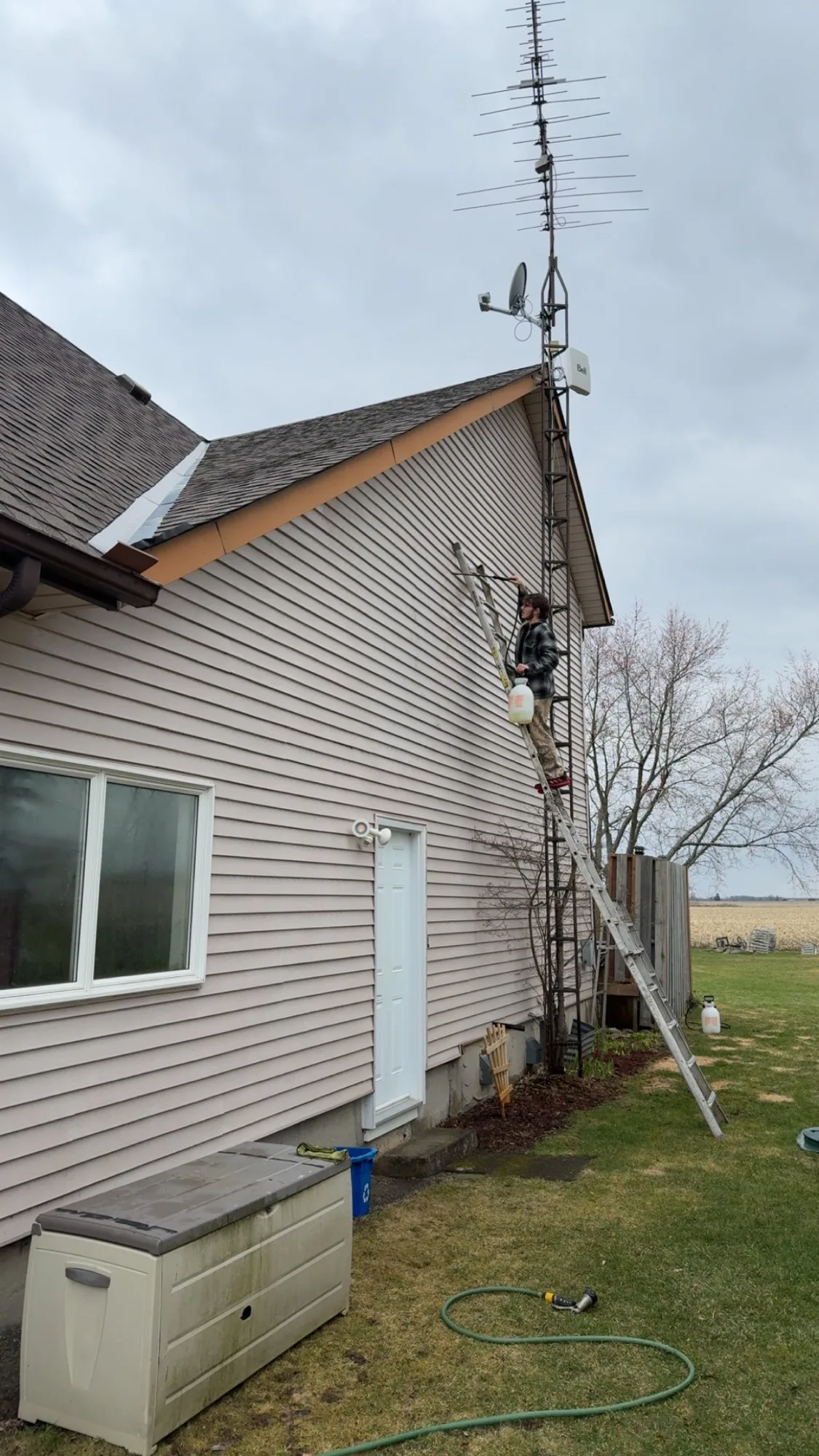 D&D Home Services technician on extension ladder soft washing tan vinyl siding on rural Kitchener farmhouse with antenna tower