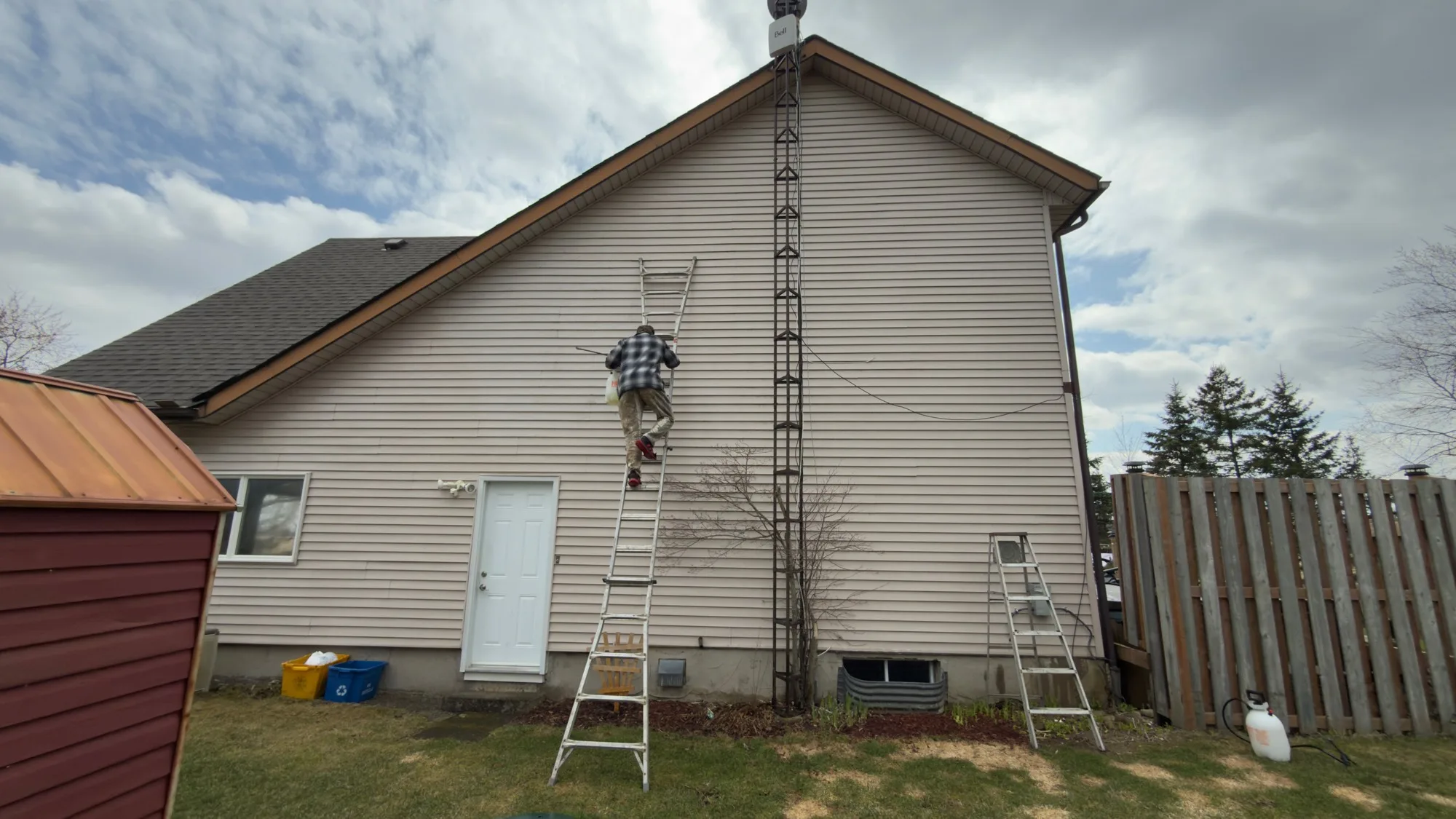 D&D Home Services technician applying soft wash solution to high gable vinyl siding on K-W rural home