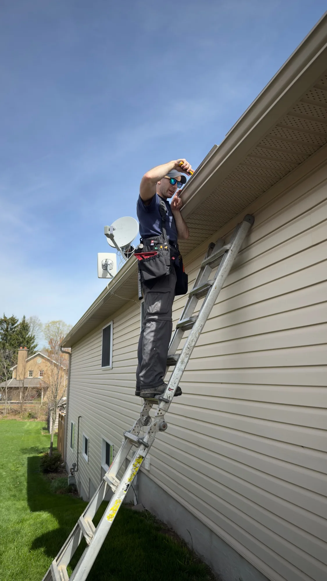 D&D Home Services technician with full tool belt measuring eavestrough run on tan vinyl home before guard install