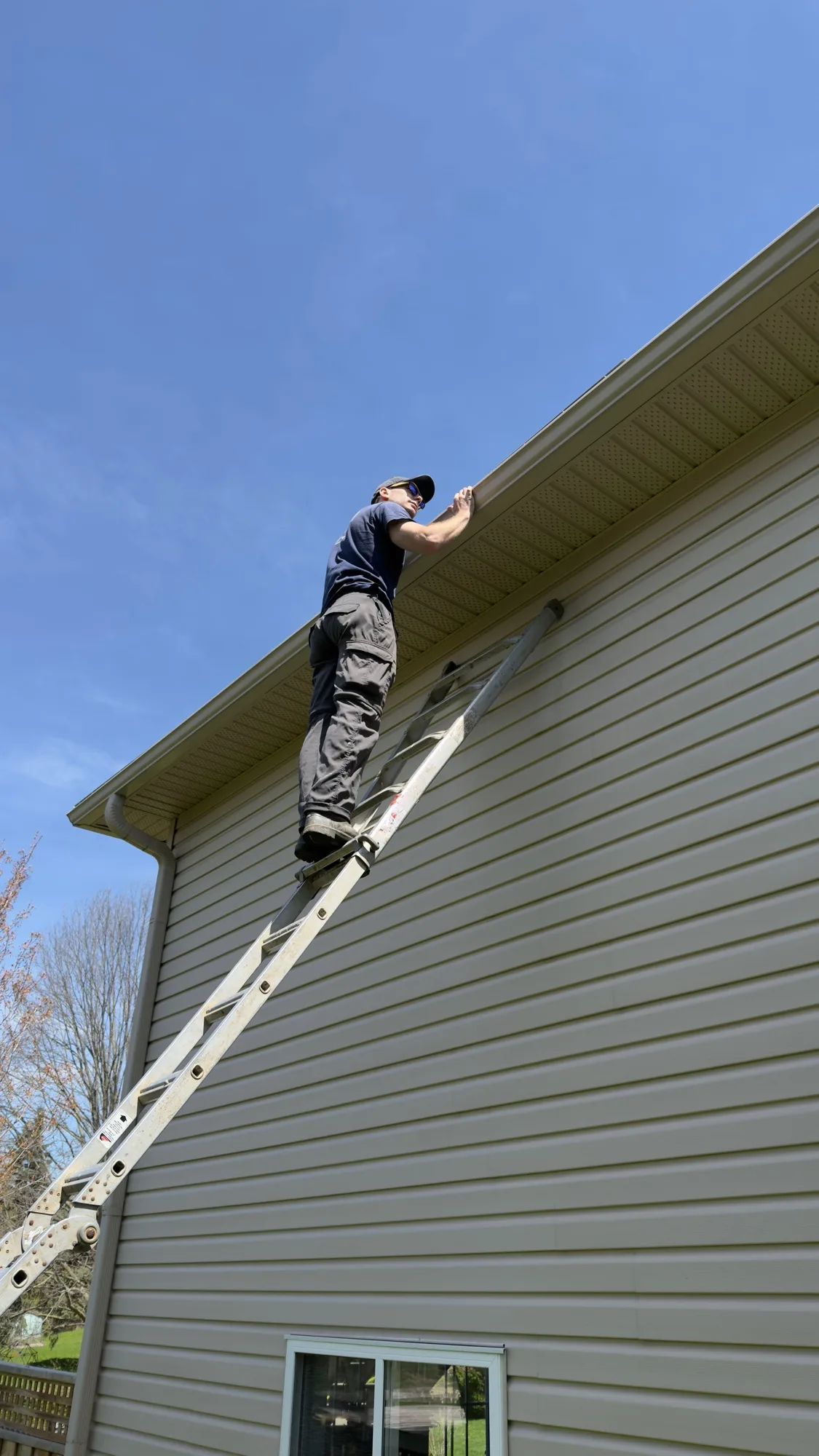 D&D Home Services technician on extension ladder reaching up at soffit line on a tan vinyl-sided two-storey home in Kitchener-Waterloo