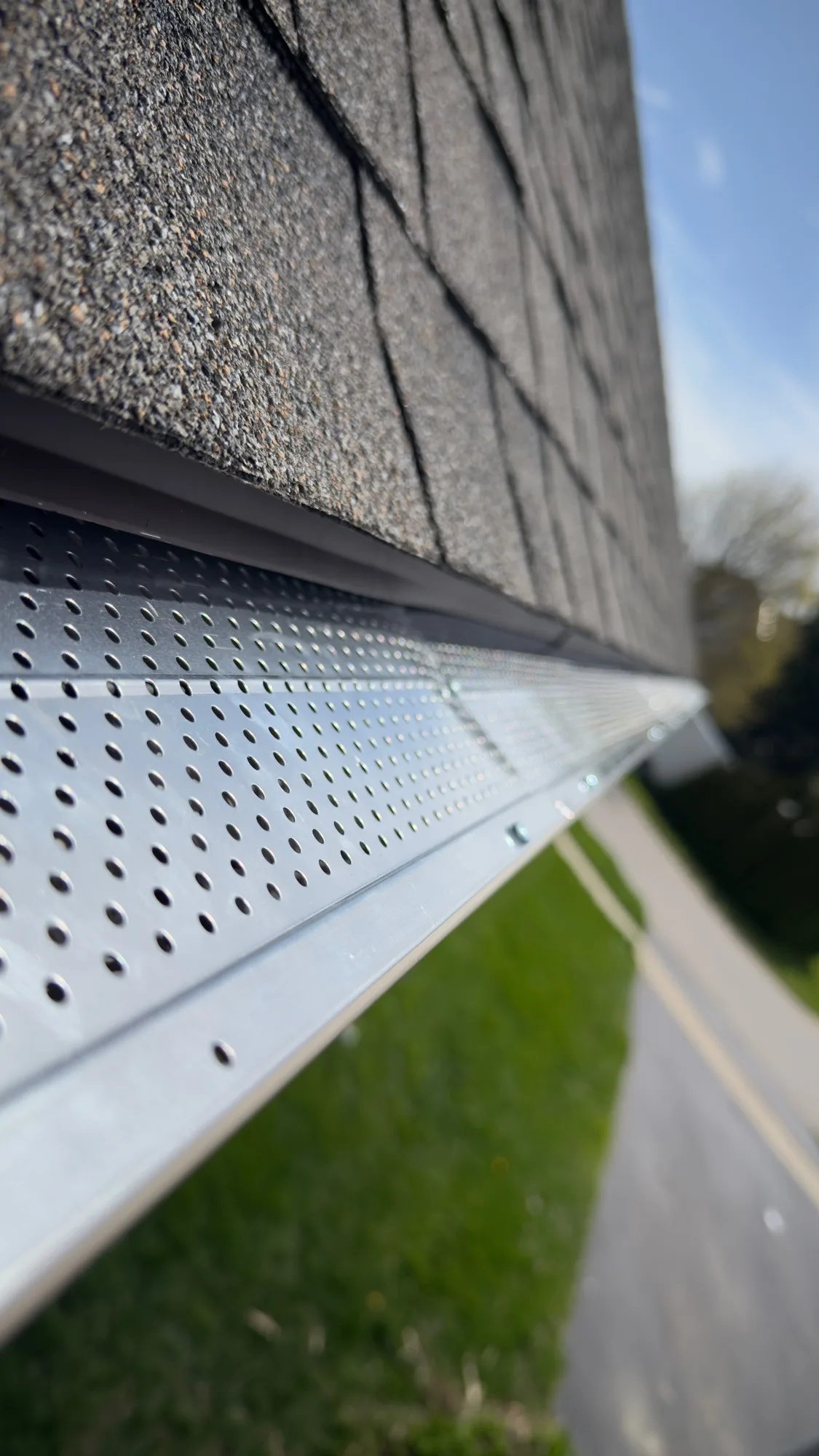 Hero close-up of silver perforated gutter guard along grey asphalt shingle roof edge with green lawn and street below — D&D Home Services Kitchener install